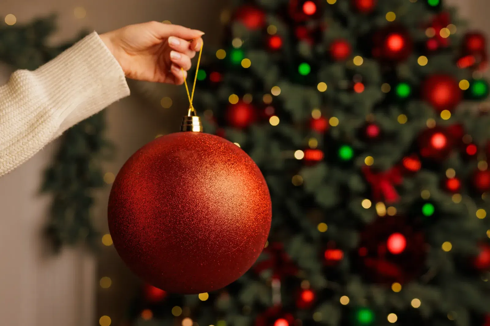 Frau hält rote große Weihnachtskugel vor einen geschmückten Christbaum.